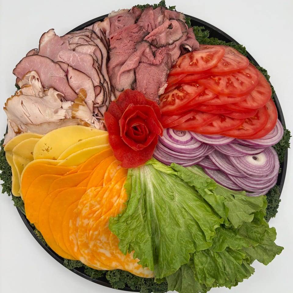 A deli platter with sliced meats, cheeses, tomatoes, red onions, and lettuce, garnished with a red pepper rose.