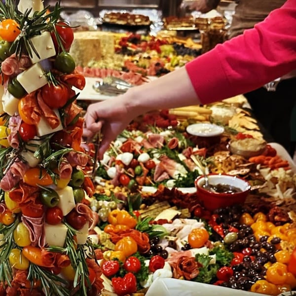 A person's hand reaching for food on a large, bountiful charcuterie board with a deli-style spread.