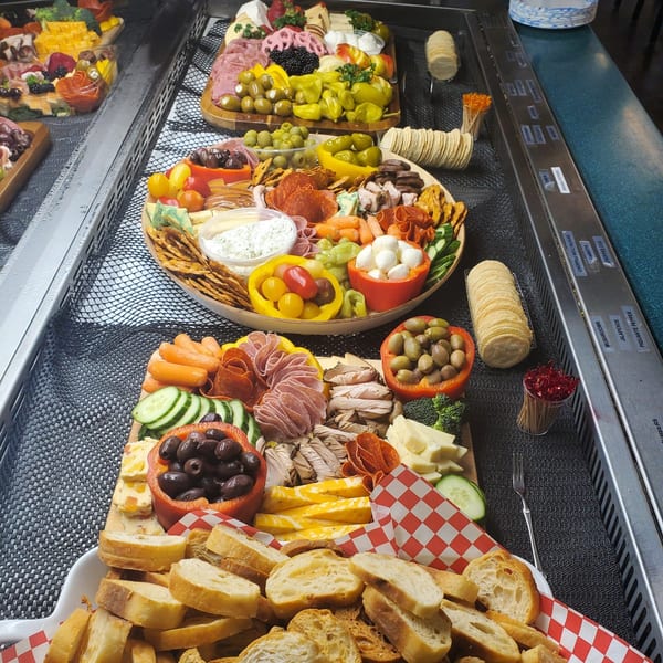 Three deli charcuterie boards overflowing with meats, cheeses, olives, and vegetables on a buffet line.