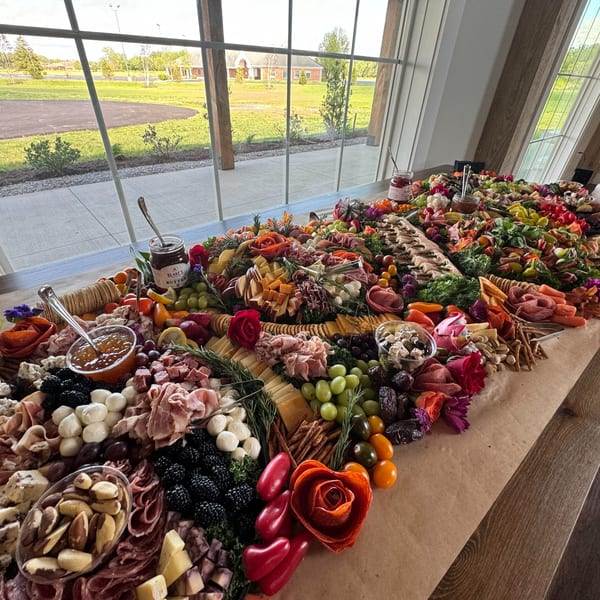 A long deli charcuterie board filled with meats, cheeses, fruits, and nuts, by a large window.