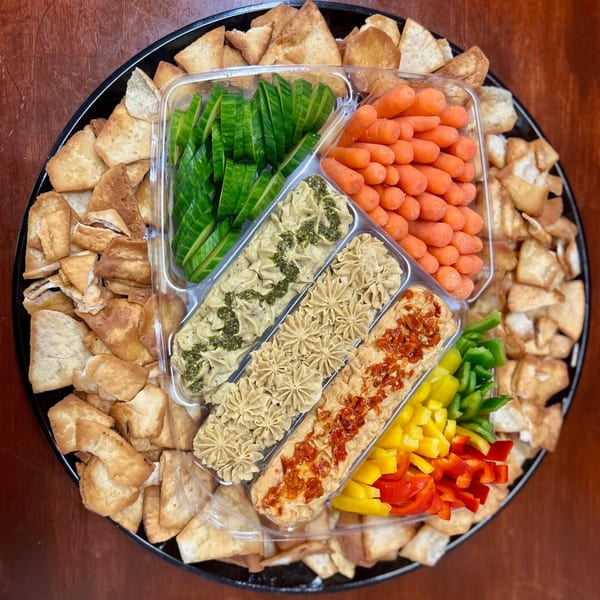 A Harvest deli platter with pita chips, sliced cucumbers, baby carrots, bell peppers, and three dips.