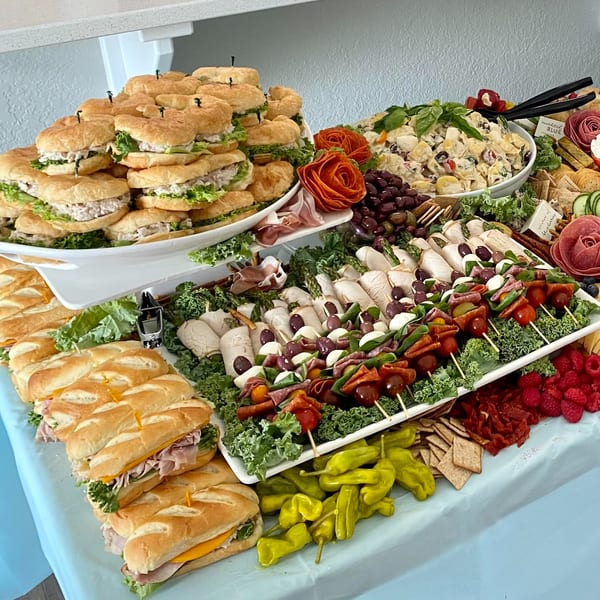 A deli spread with sandwiches, pasta salad, skewers, and charcuterie on a light blue tablecloth.
