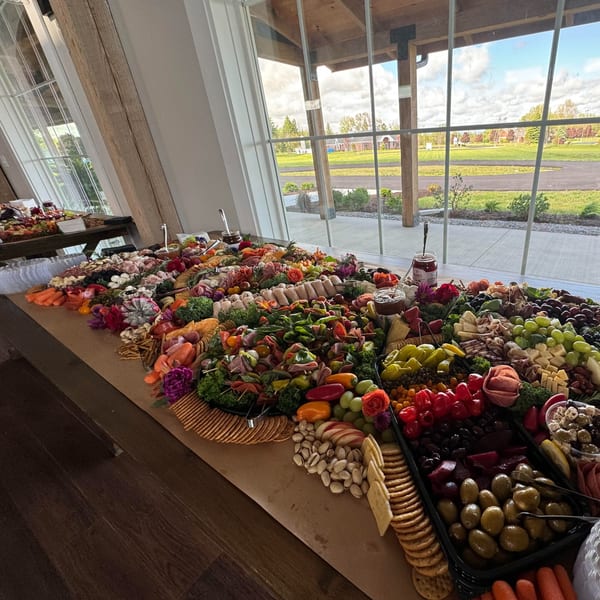 A deli charcuterie board overflowing with meats, cheeses, fruits, and vegetables by a large window.